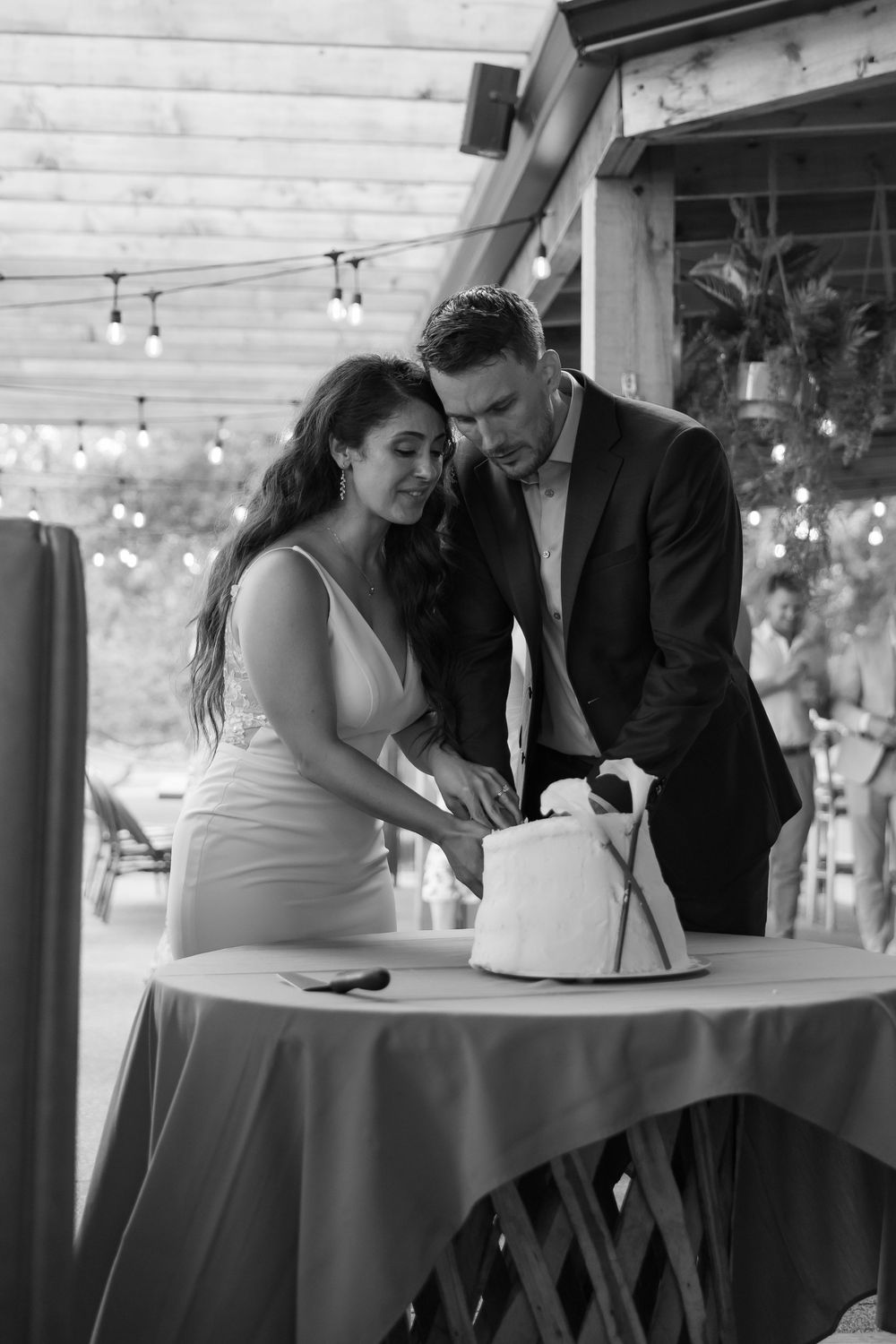 A couple in formal attire cut their wedding cake together at an outdoor reception with string lights in the background.
