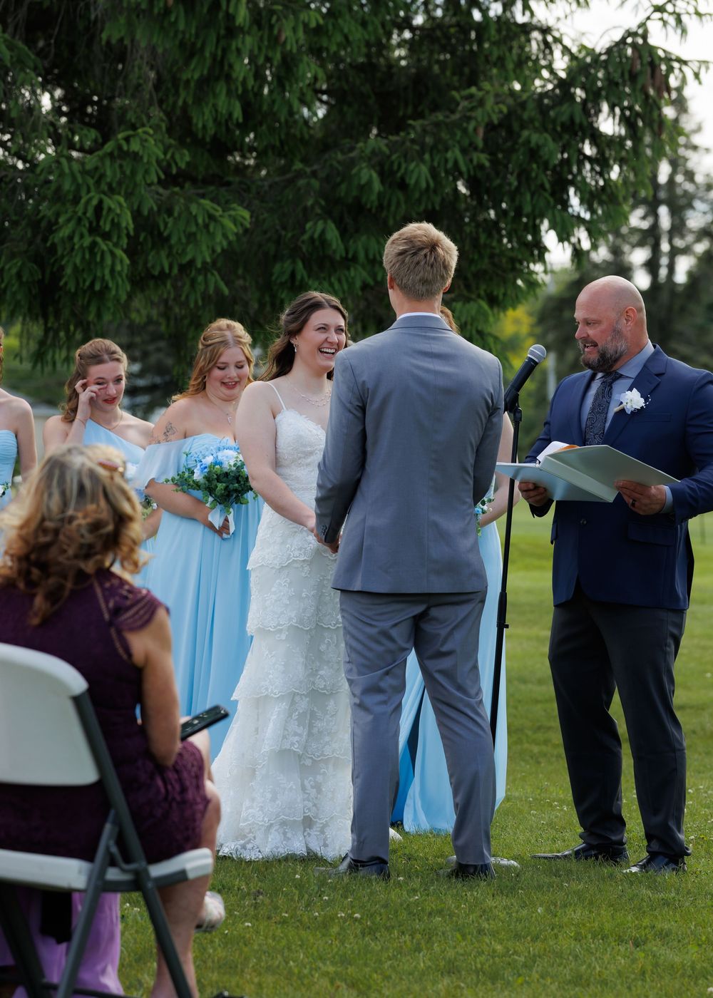 A wedding party gathers outdoors on green grass, with bridesmaids in light blue dresses and groomsmen in navy suits.