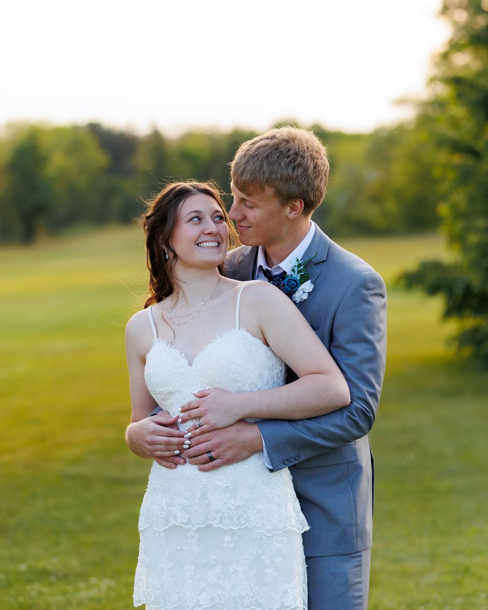 Newly married couple embraces on a grassy hillside during golden hour sunset in their elegant wedding attire.