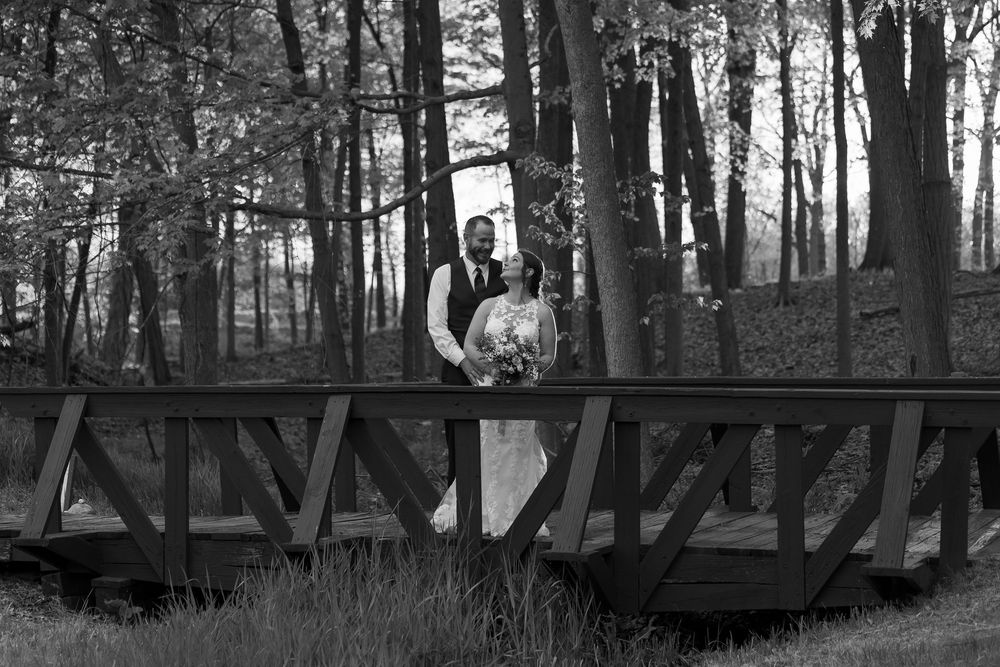 Black and white photo of a bride and groom standing on a wooden bridge surrounded by trees.