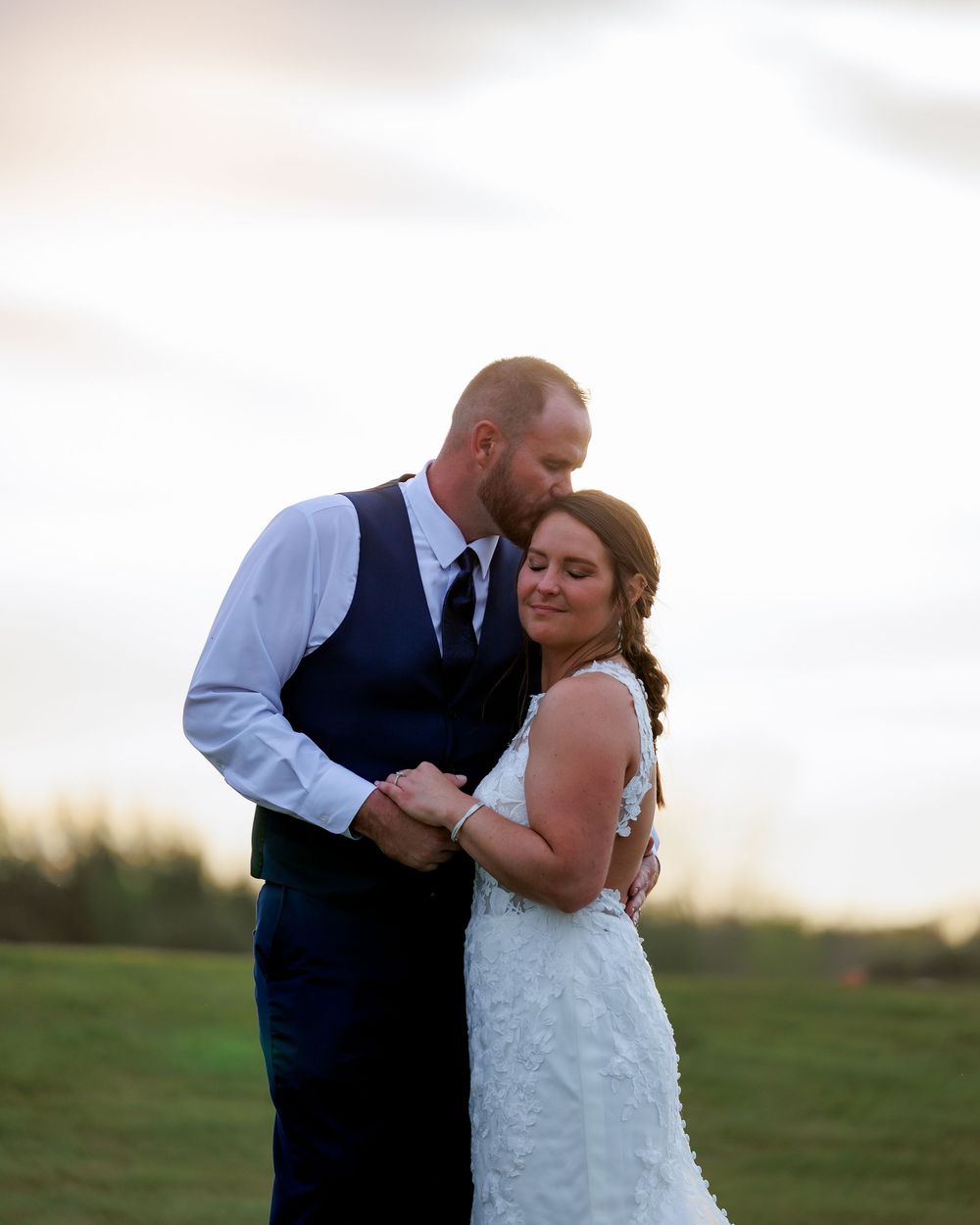 Newlywed couple embracing at golden hour in a field, bride in white lace gown and groom in navy vest and tie.