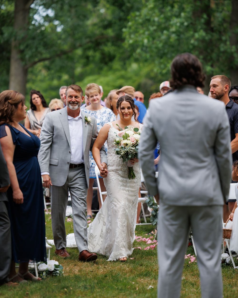 Bride walks down outdoor aisle with father, holding white bouquet, as groom waits in gray suit at garden wedding ceremony.