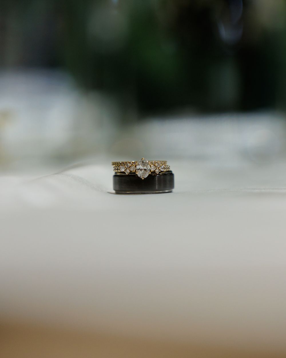 Gold diamond engagement ring stacked on a dark wedding band displayed on a white surface.