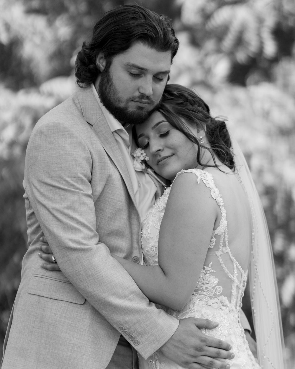 Black and white wedding photo of bride and groom embracing tenderly outdoors in elegant attire.