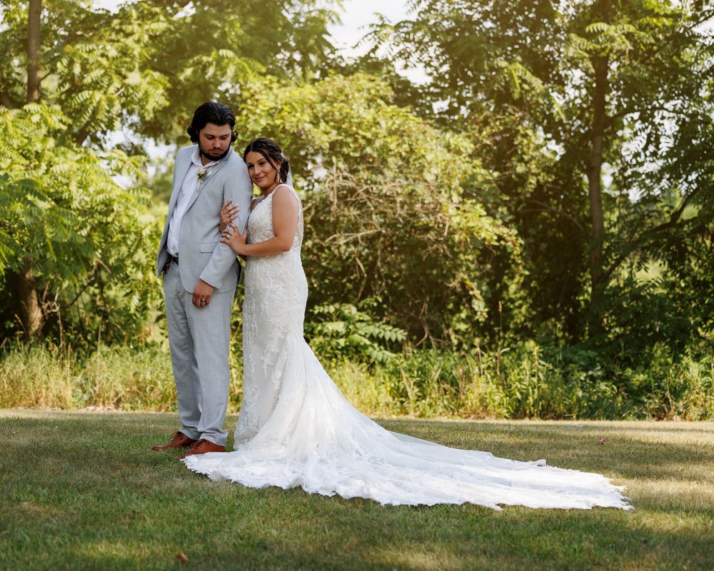 A bride and groom pose outdoors on green grass surrounded by lush trees on their wedding day.