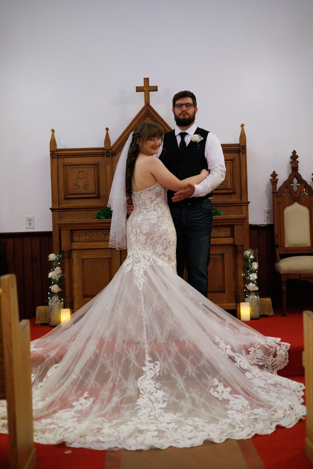 A bride in a flowing lace wedding gown with a dramatic train poses with her partner in a church setting.