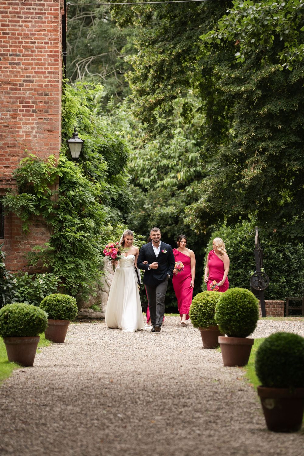 Members of a wedding party walk along a gravel path bordered by topiary plants and brick pillars at a garden venue.