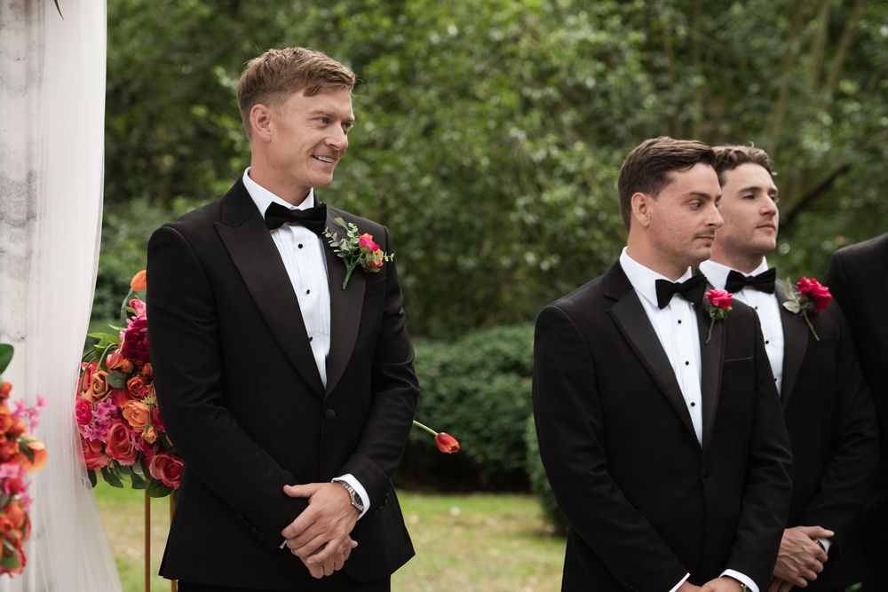 A group of groomsmen in black tuxedos standing together outdoors at a formal wedding ceremony.