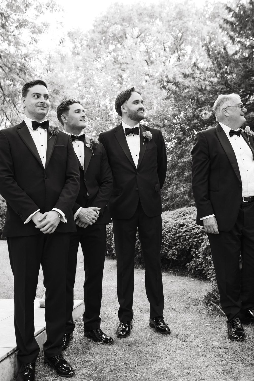 Black and white panoramic photo showing groomsmen standing in a row wearing tuxedos outdoors during a wedding.