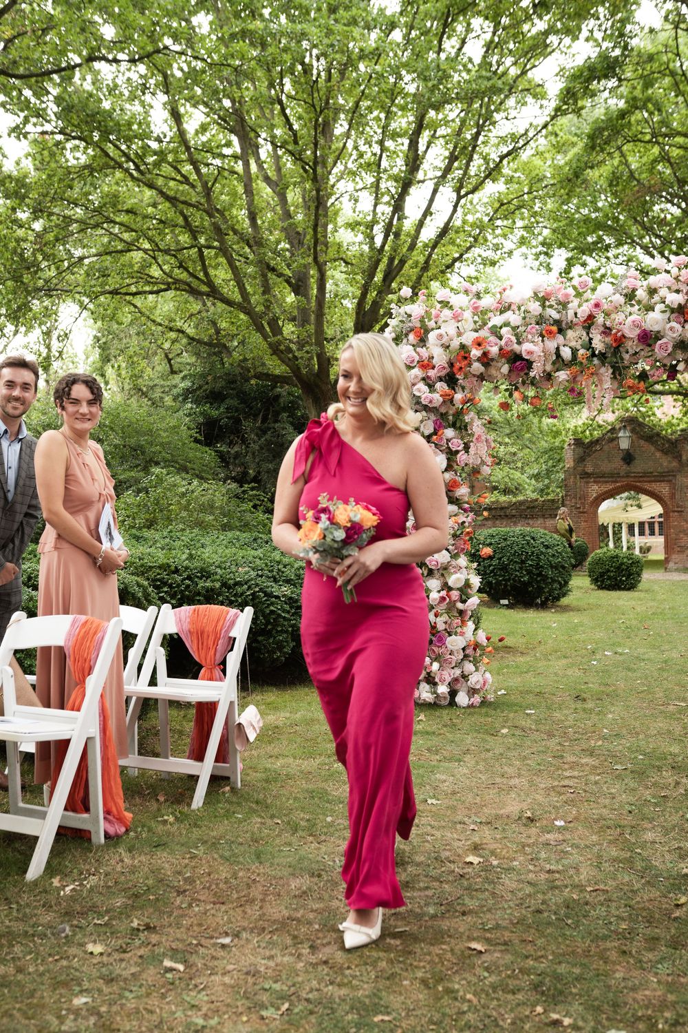 A bridesmaid in a vibrant fuchsia one-shoulder gown walks down an outdoor wedding aisle decorated with pink flowers.