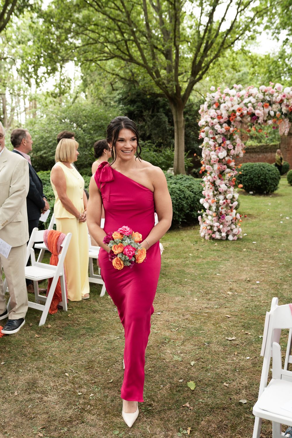 Outdoor wedding scene with people in formal attire gathered near white chairs and floral decorations in a garden setting.
