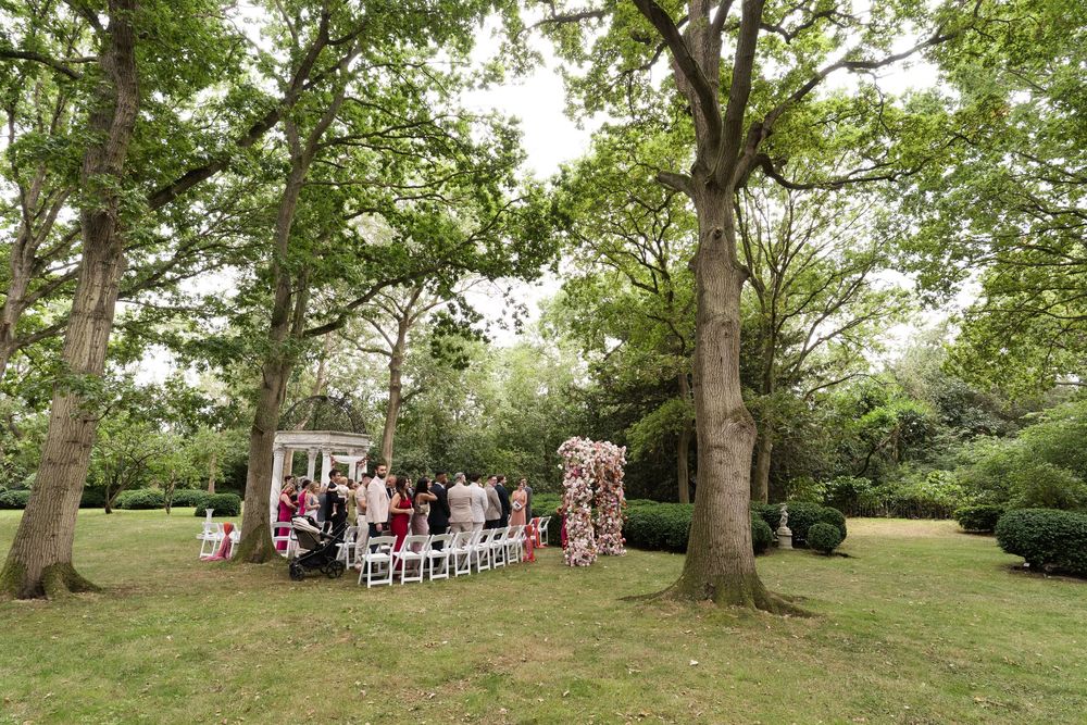 A panoramic view of guests seated at an outdoor wedding ceremony among tall trees in a lush green garden setting.