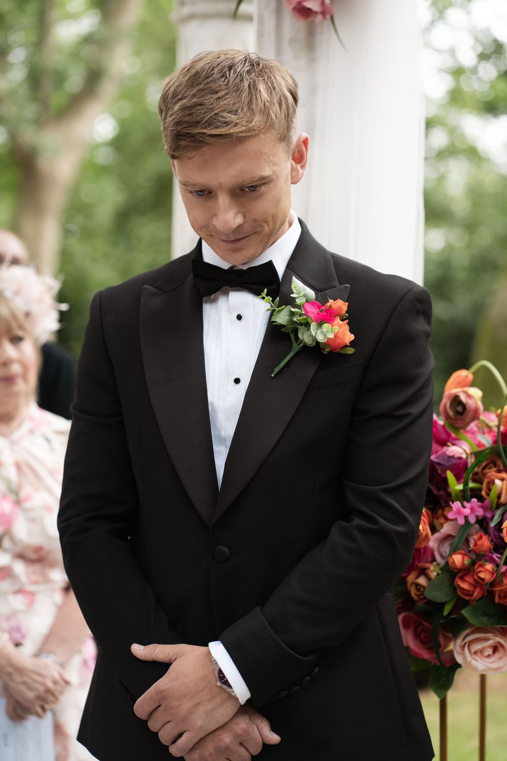 A groom in a black tuxedo and coral boutonniere looks down during an outdoor wedding ceremony.