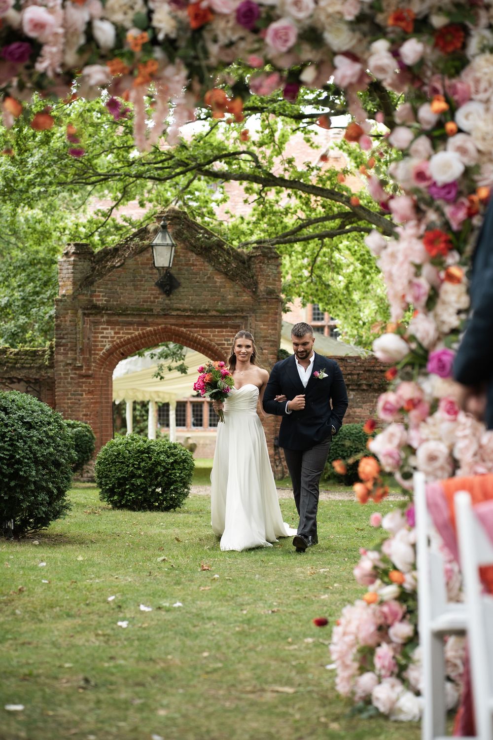 A wedding ceremony setup with a beautiful floral arch over a brick entrance and lush greenery in a garden setting.