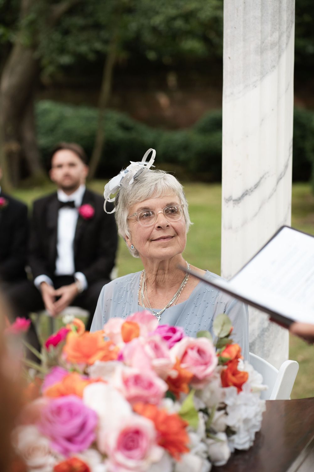 An elderly woman with glasses and a joyful expression sits near colorful pink and orange flowers at an outdoor event.