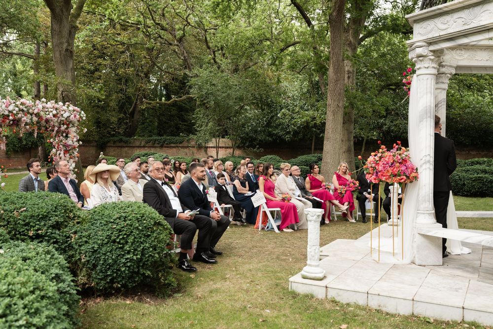 An outdoor wedding ceremony takes place in a lush garden setting with white columns and guests seated on lawn chairs in rows.