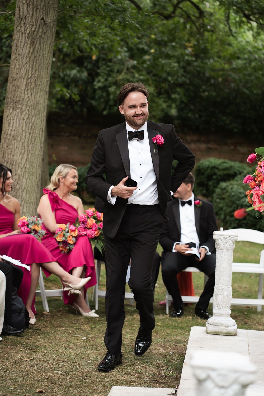 Wedding party in formal attire sits and laughs together on white chairs outdoors beneath tall trees.