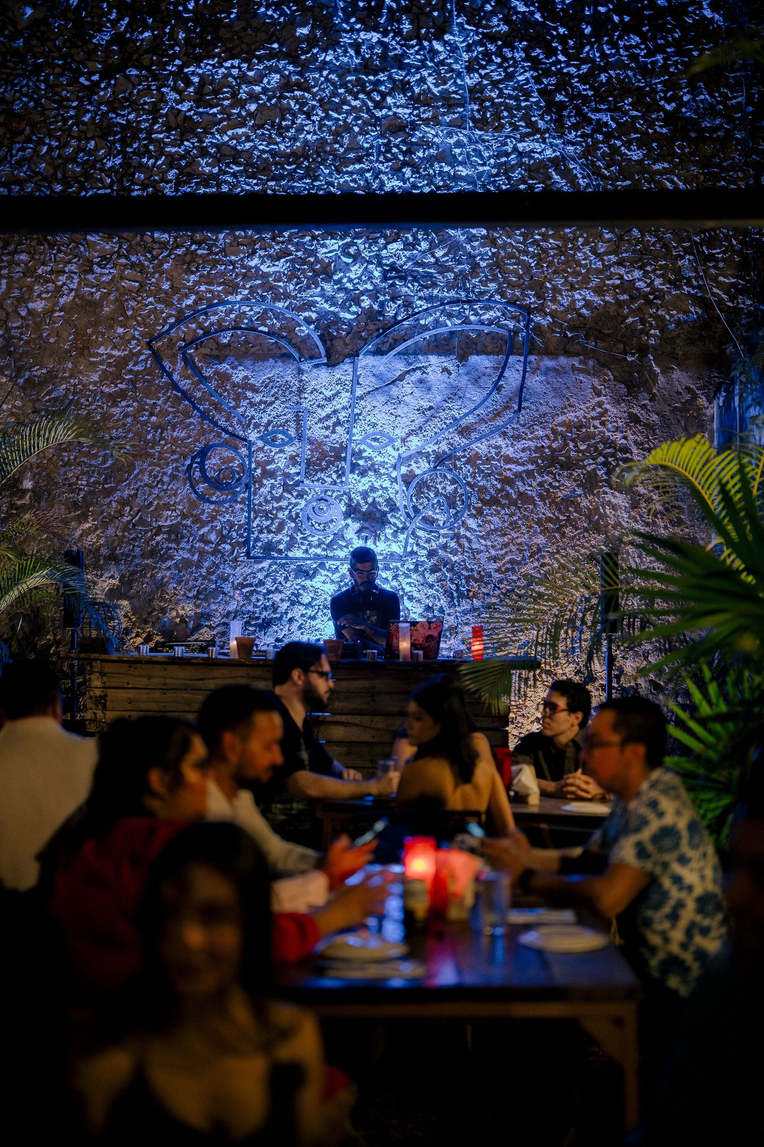 A dimly lit restaurant interior with blue lighting on textured walls, silhouetted diners seated at tables enjoying their meals.