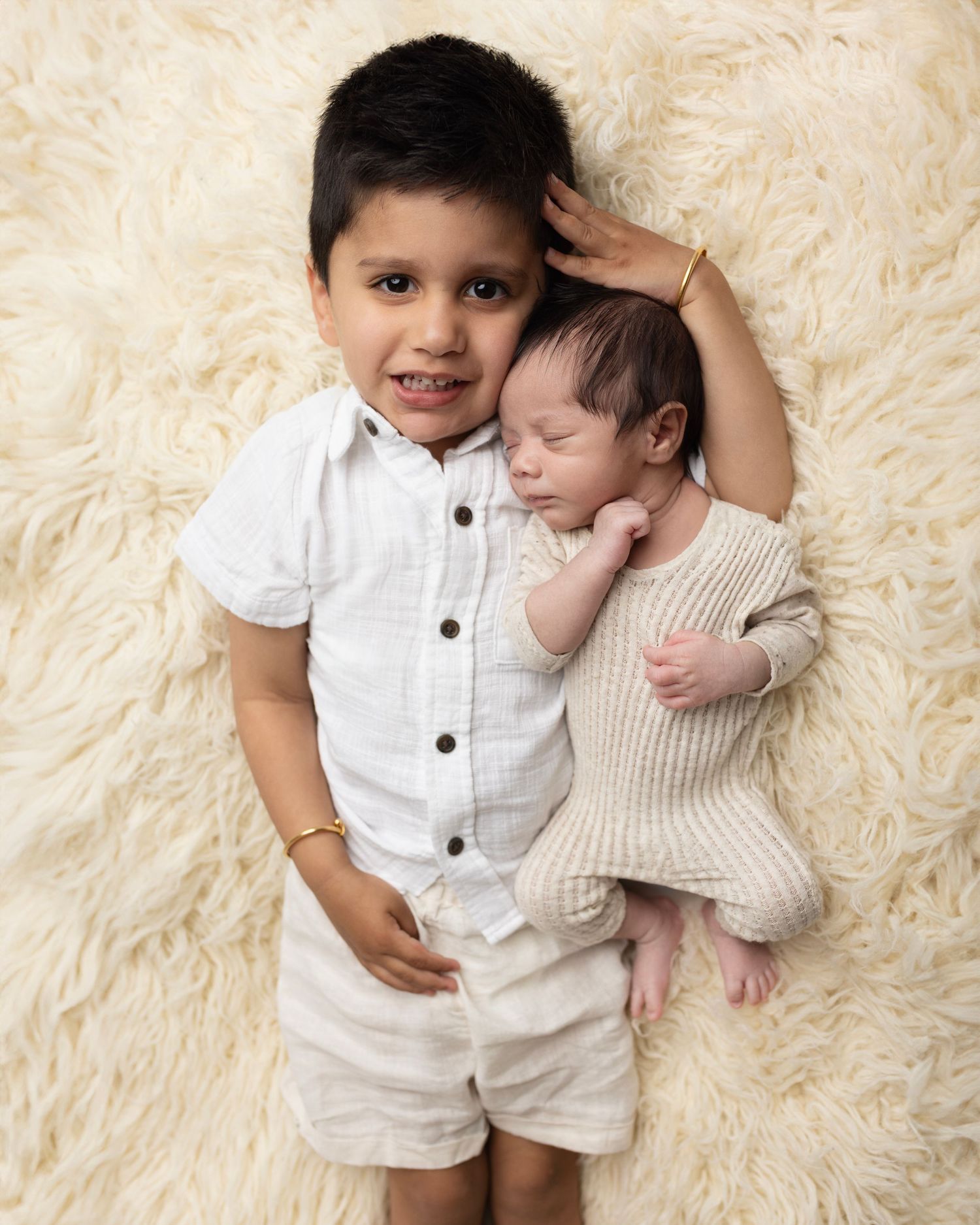 Two small children in white outfits lying together on a cream-colored fluffy rug during a portrait photography session.