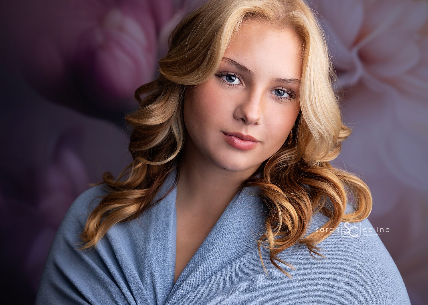 A portrait of a model wearing a blue sweater against a purple backdrop with soft lighting and wavy blonde hair.