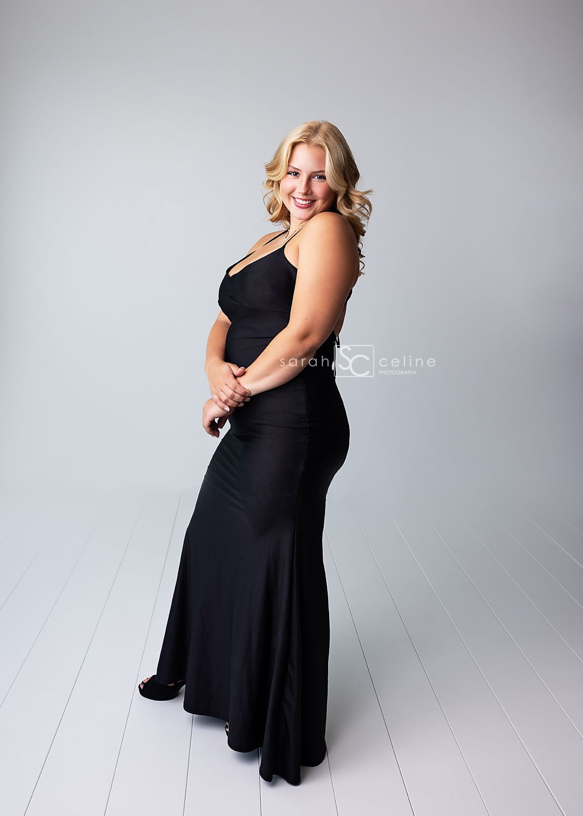A person in an elegant black floor-length evening gown poses gracefully against a light gray studio backdrop.
