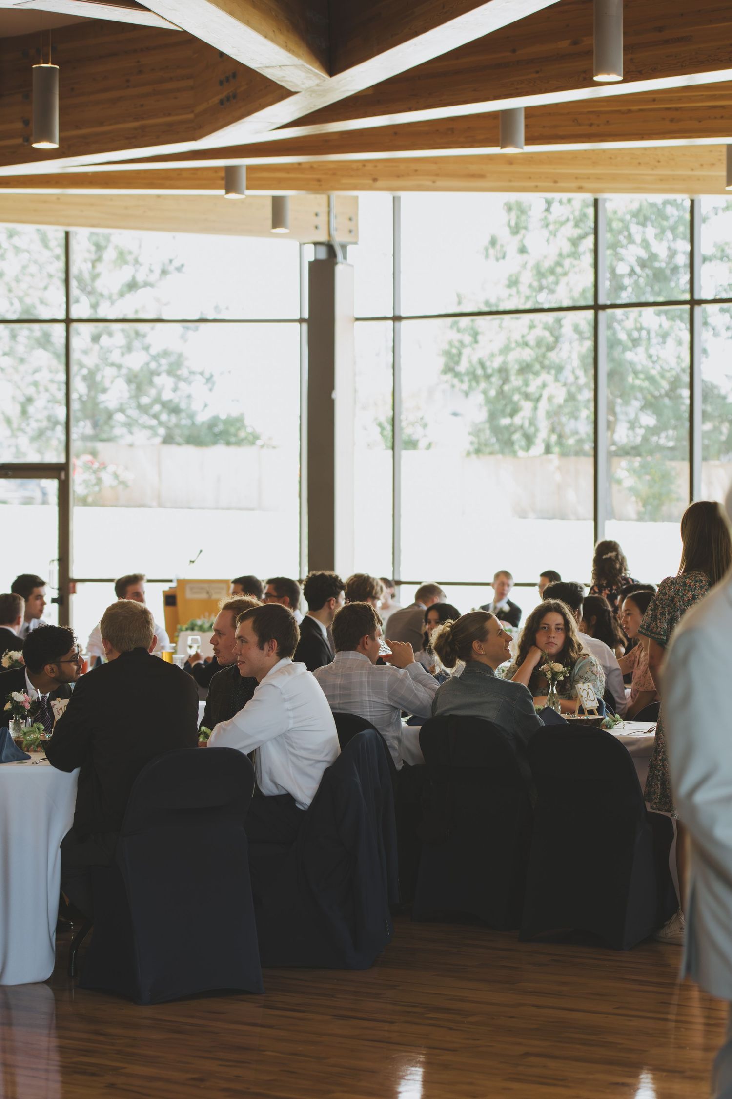 A crowded event venue with wood floors and tall windows shows attendees seated at tables during a formal gathering.