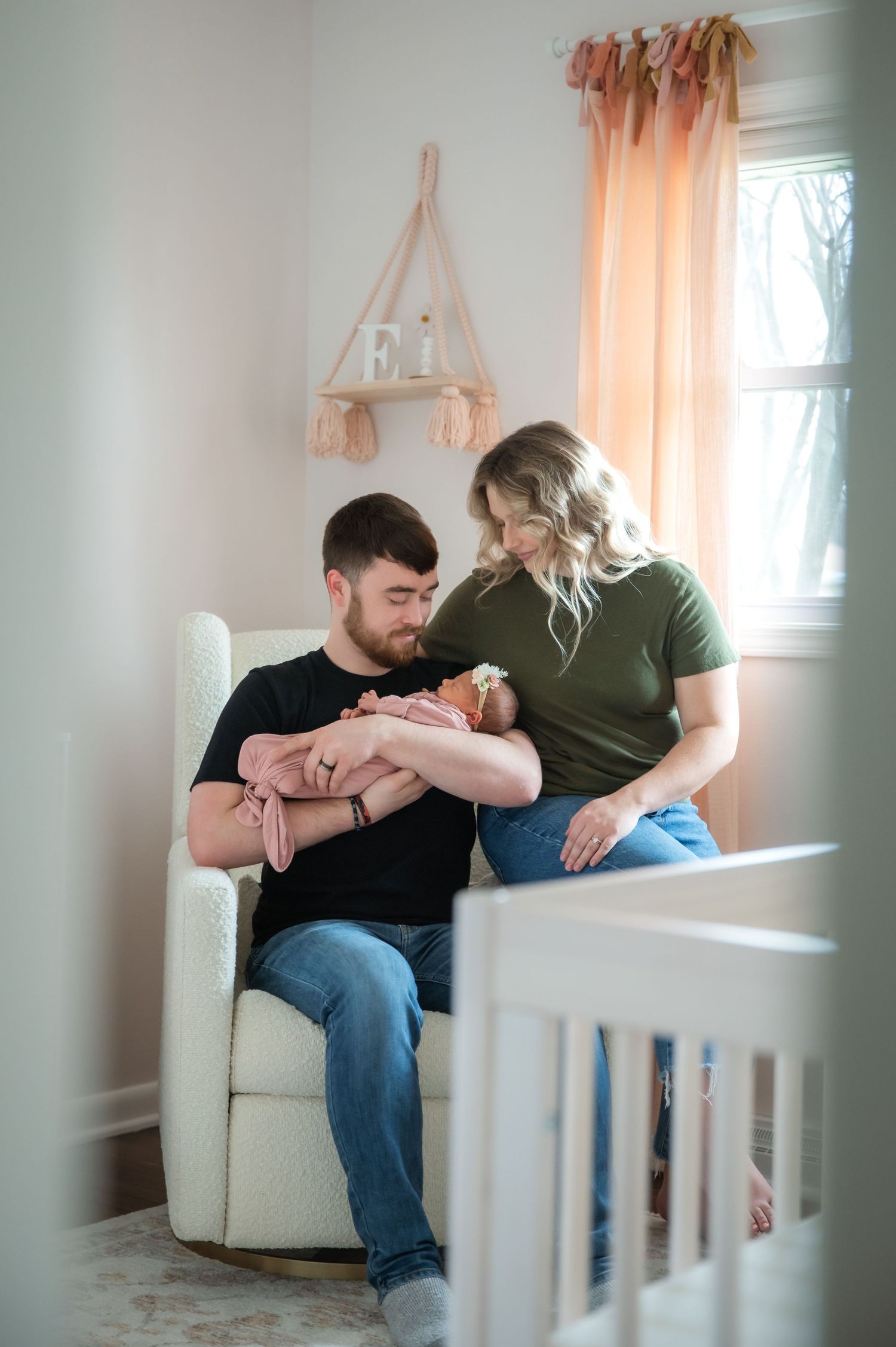A couple sits together in a white chair by a window with soft peachy curtains in a baby's nursery.
