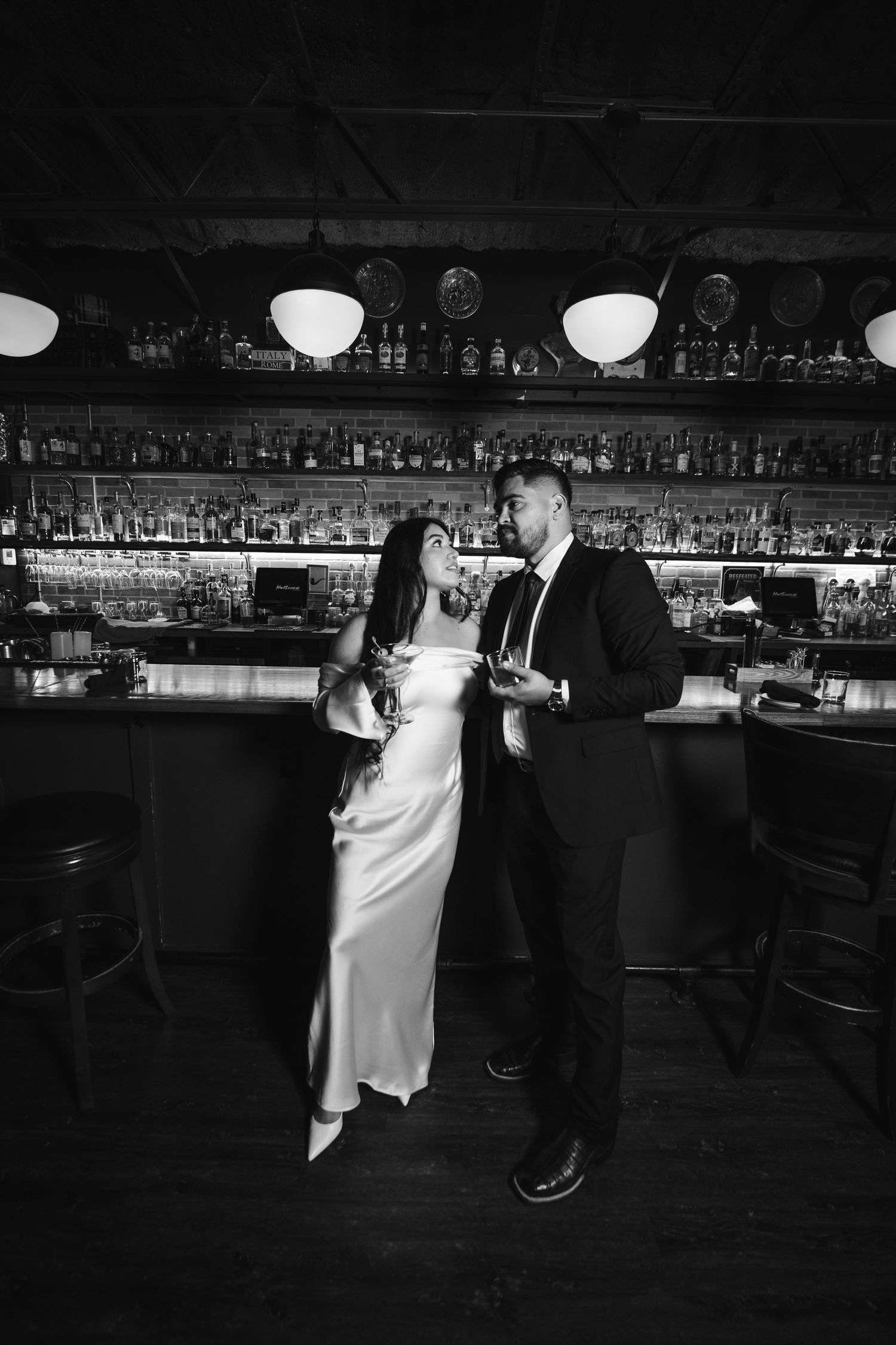 A romantic dance scene unfolds in a dark bar with bottles lining the shelves in the background.