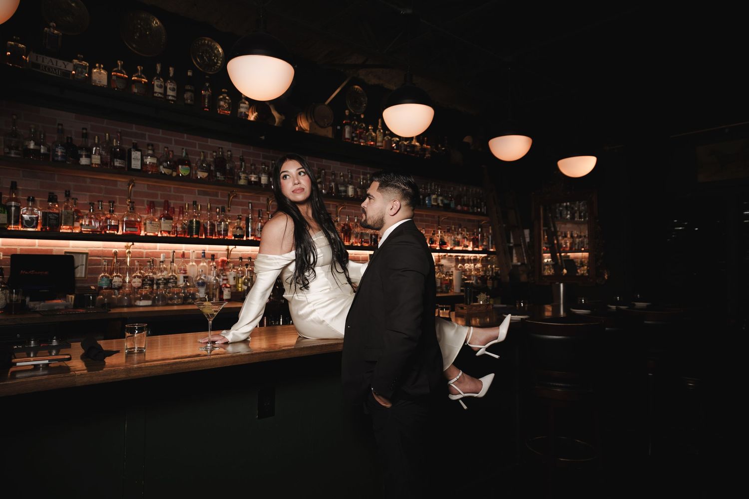 A couple shares a playful dance moment in a moody bar setting with warm lighting.
