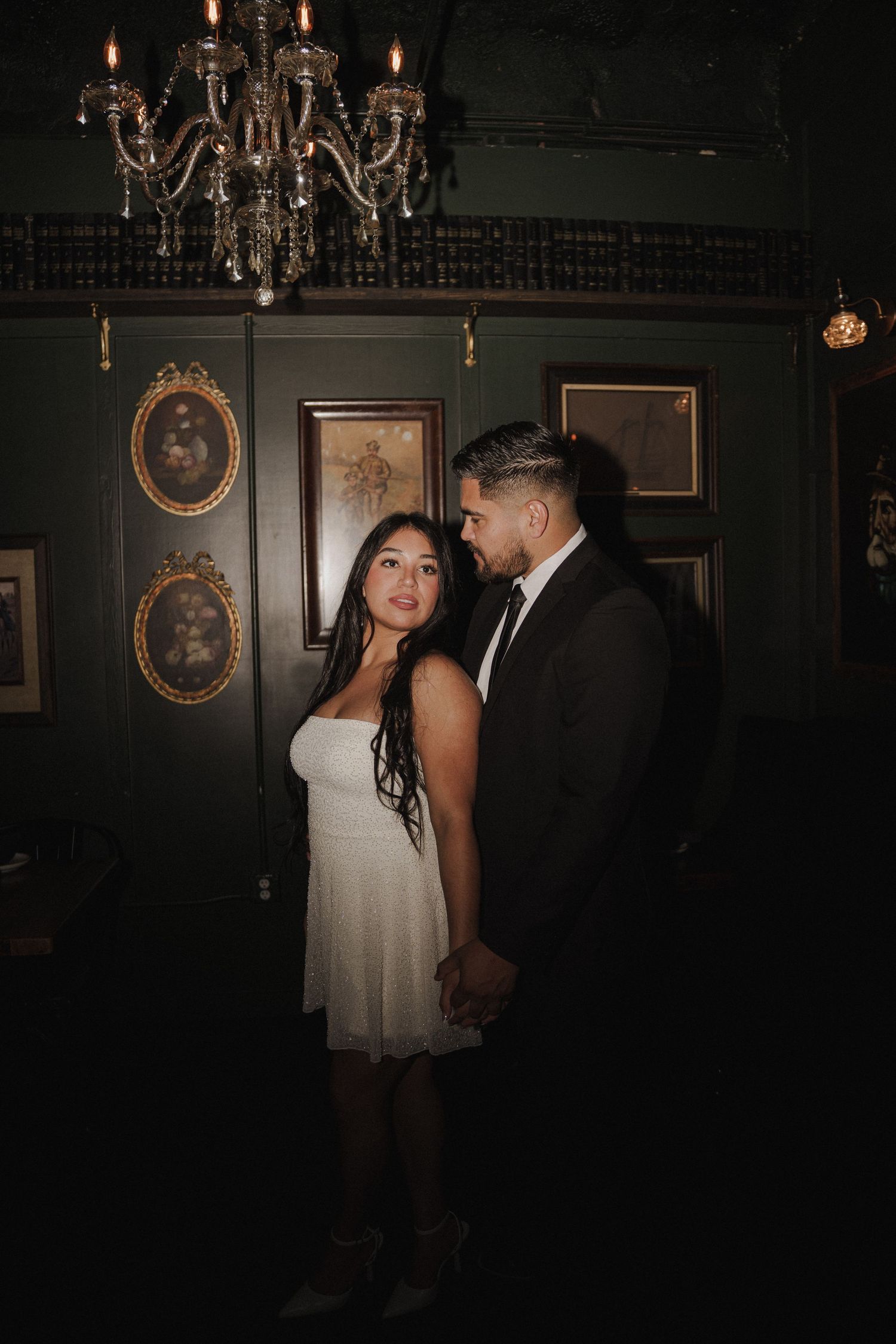 A moody bar scene with ornate chandeliers and vintage portraits on dark walls.