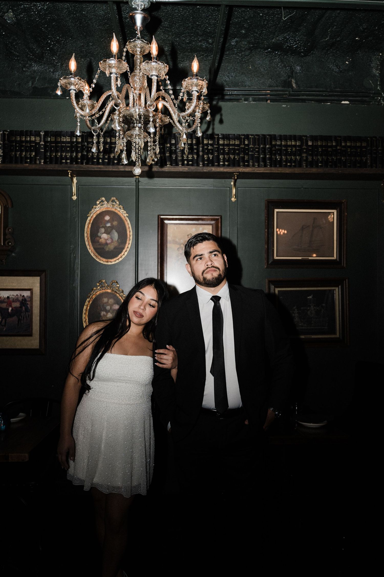 A couple poses beneath crystal chandeliers in a vintage-styled bar with dark walls.