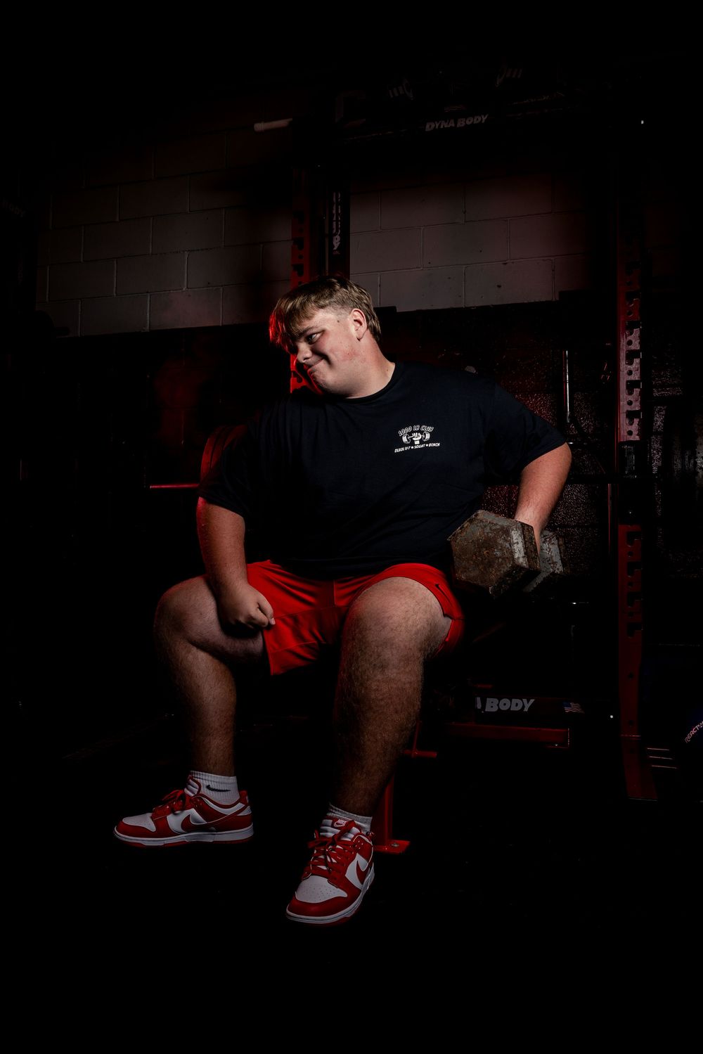 Amateur athlete in red shorts and black t-shirt sitting on weight bench with dramatic dark lighting in gym setting.