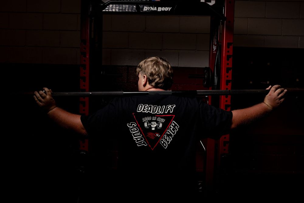 Back view of a weightlifter in a black t-shirt with text standing at a power rack in a dark gym setting.