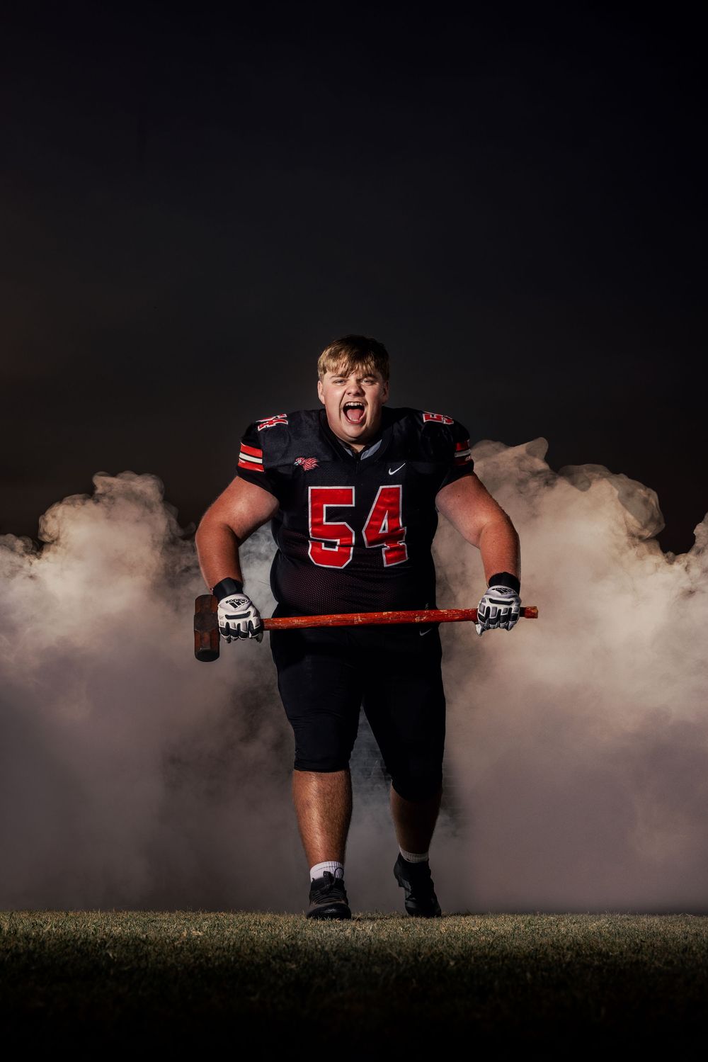 Football player in uniform number 54 lifting weights with smoke effects in dramatic outdoor photo.