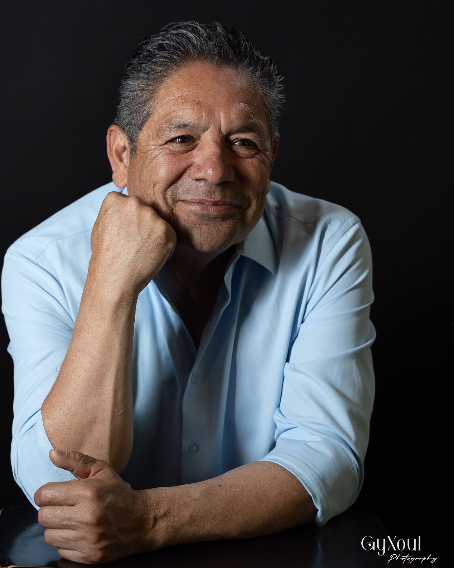 Portrait of someone in a light blue button-down shirt resting their chin on hand against dark background.