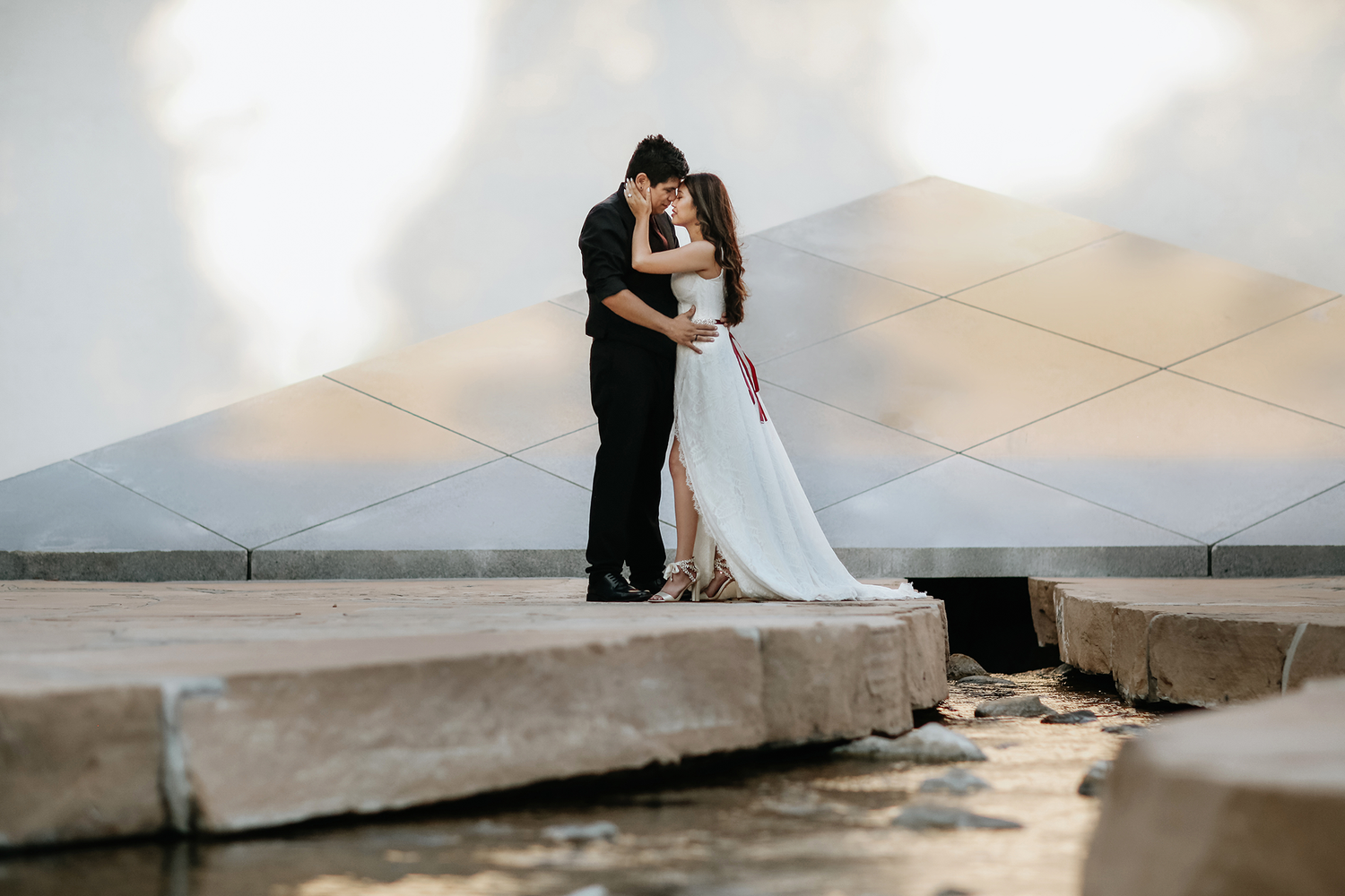 A couple shares an intimate embrace on an outdoor stone ledge with ocean views at sunset.