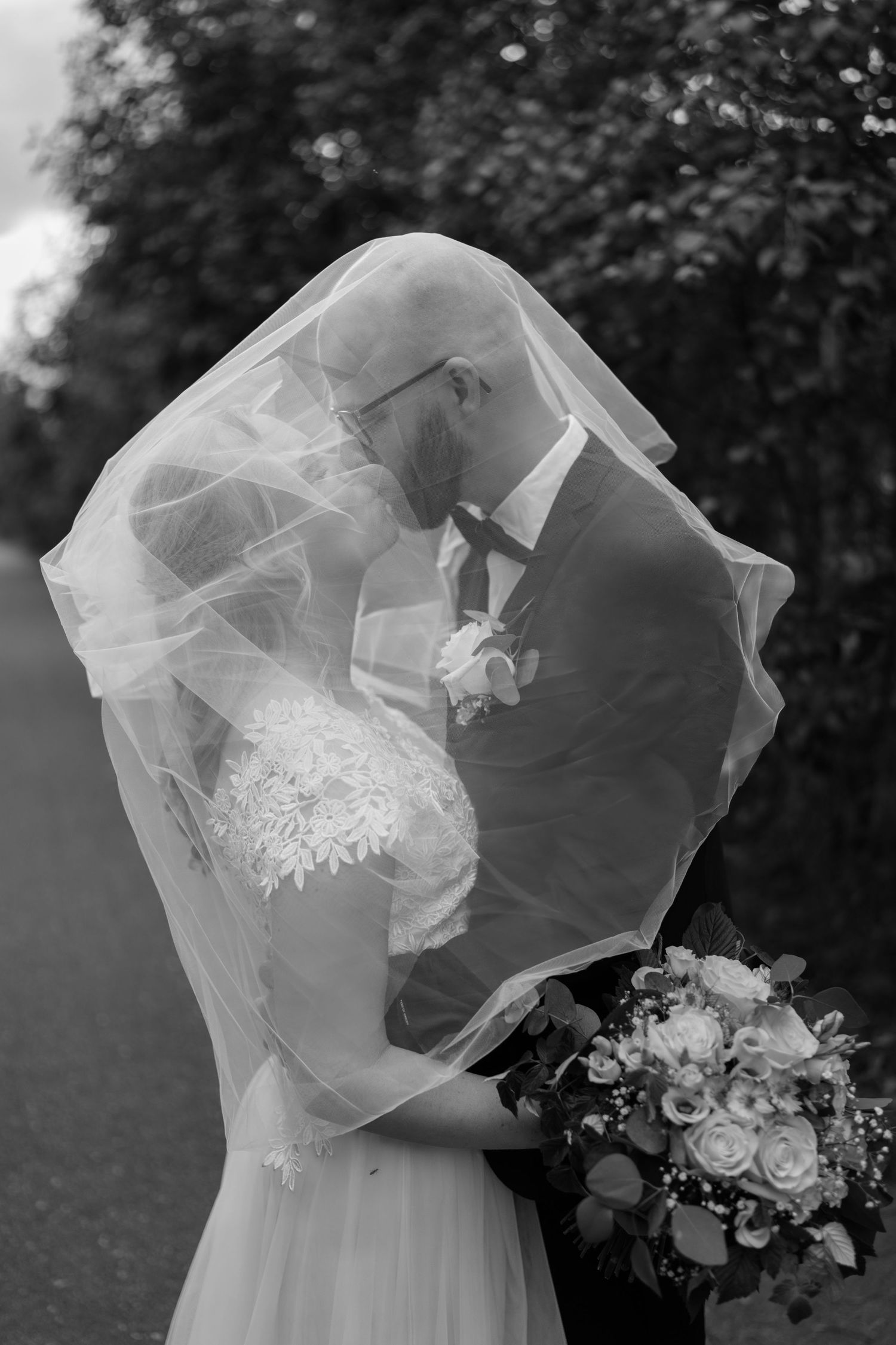 Black and white photo of bride and groom kissing under wedding veil, bride holding bouquet of roses outdoors.