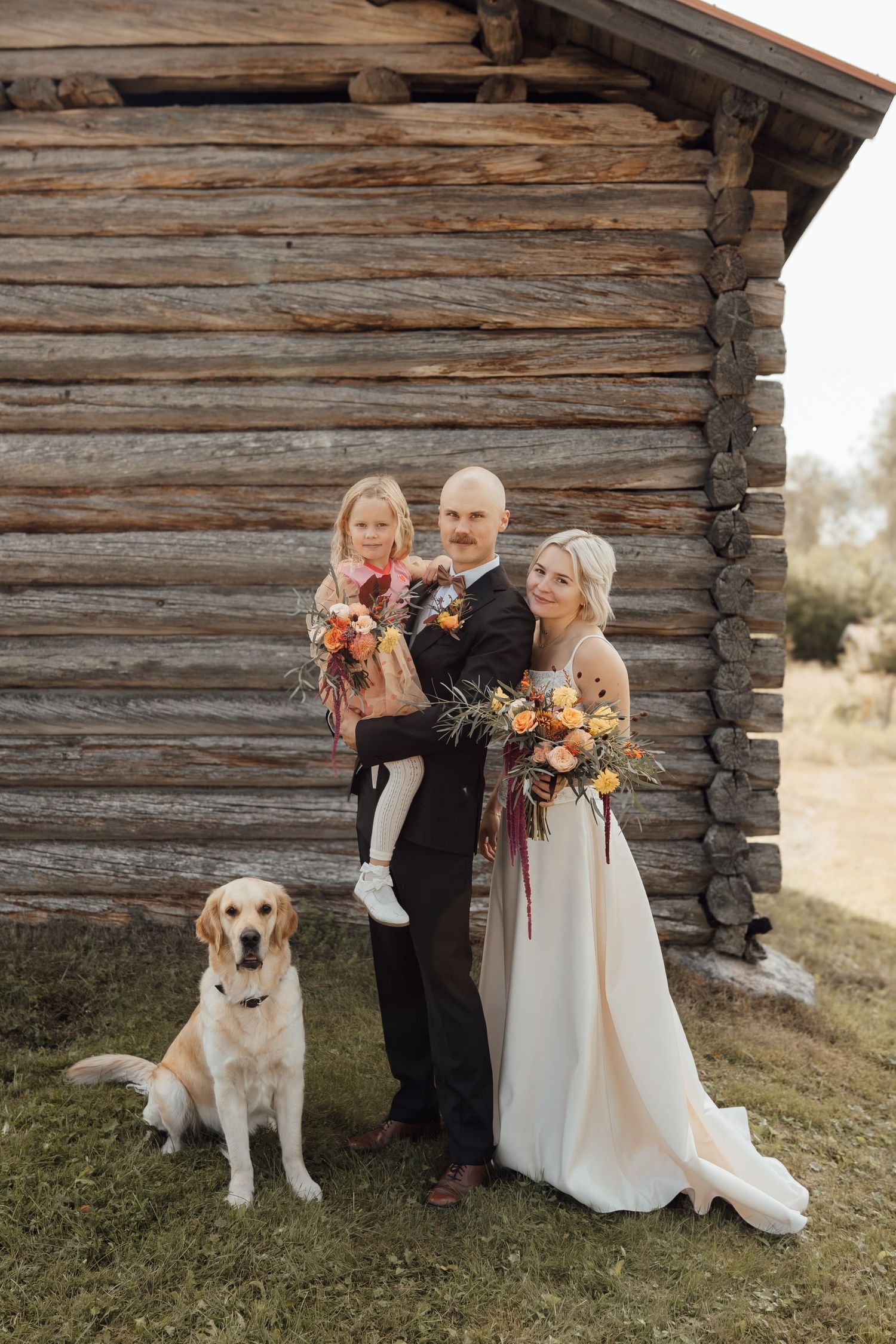 Wedding couple with child and golden retriever dog posing in front of rustic log cabin, holding colorful floral bouquets.