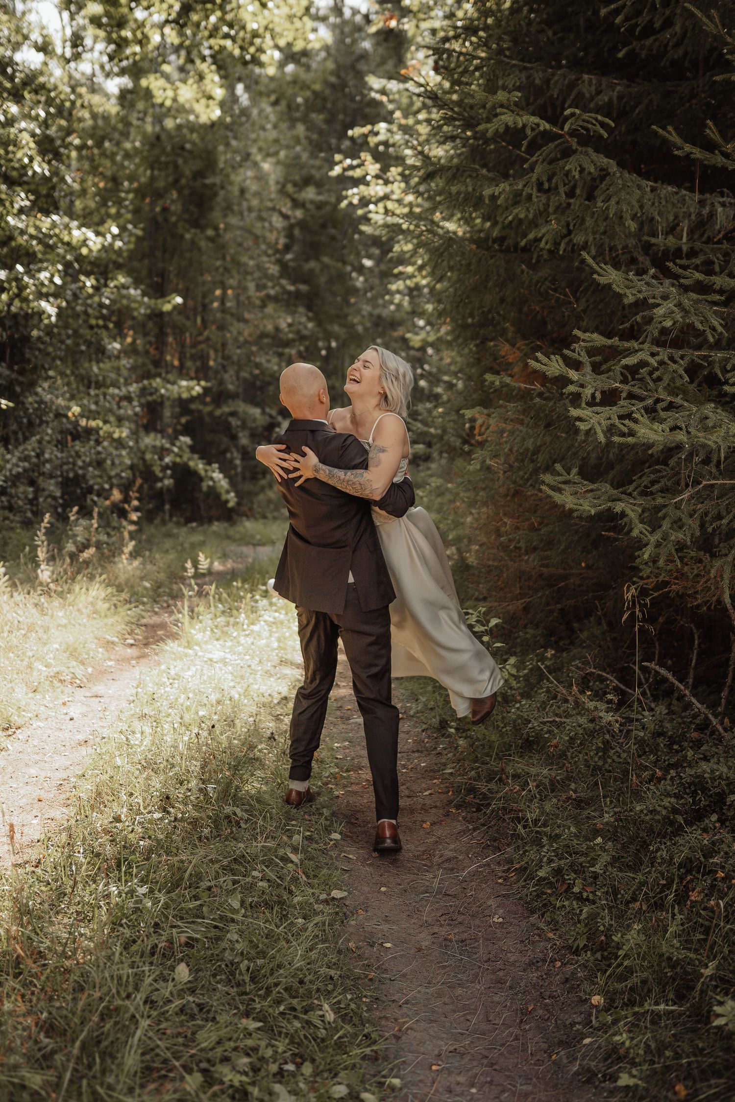A joyful couple embraces on a sunlit forest path, the groom lifting his laughing bride in a romantic outdoor wedding setting.