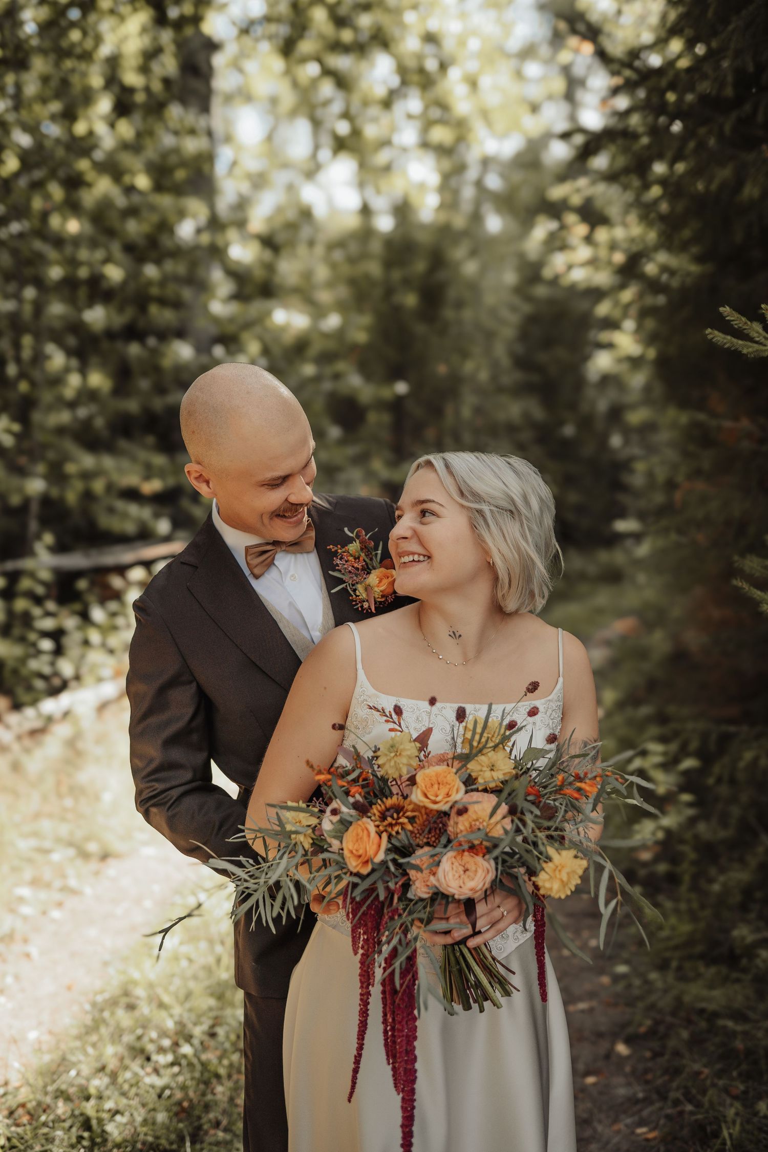 Smiling bride holding autumn bouquet with orange flowers stands with groom in brown suit outdoors in forest setting.