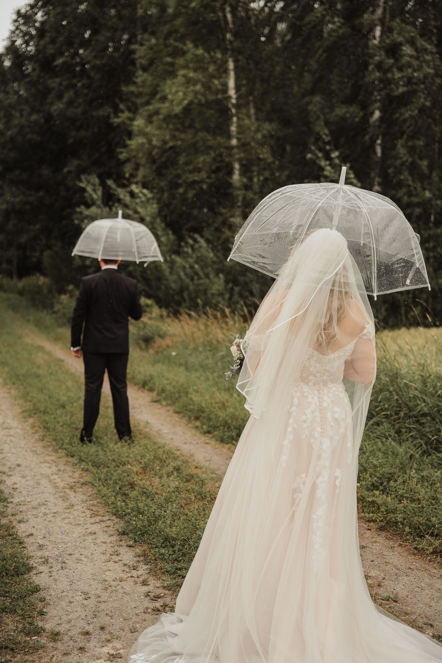 Bride and groom holding clear bubble umbrellas on a dirt path surrounded by lush greenery on a rainy wedding day.