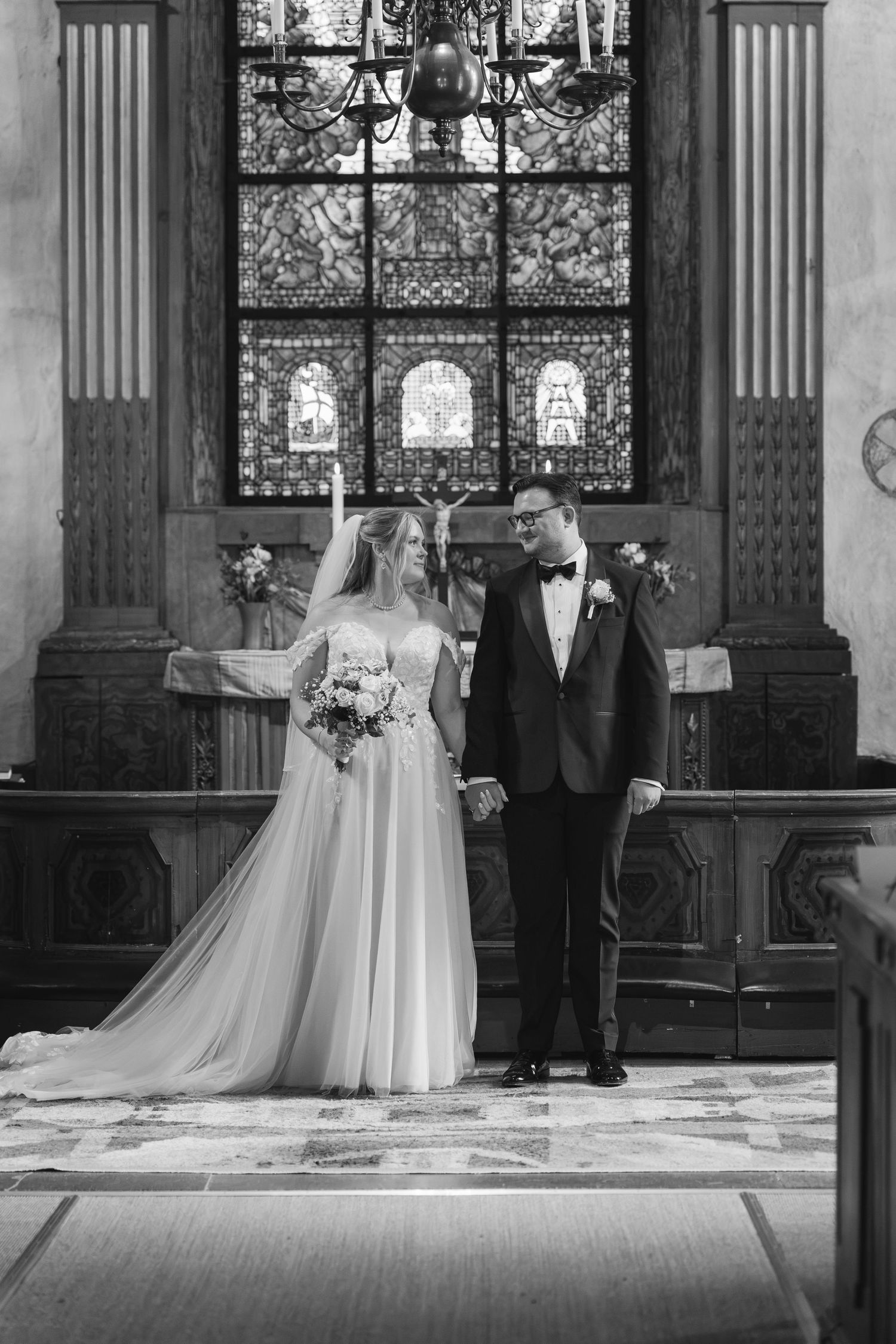Black and white photo of a bride and groom holding hands at the altar of a church with stained glass windows.
