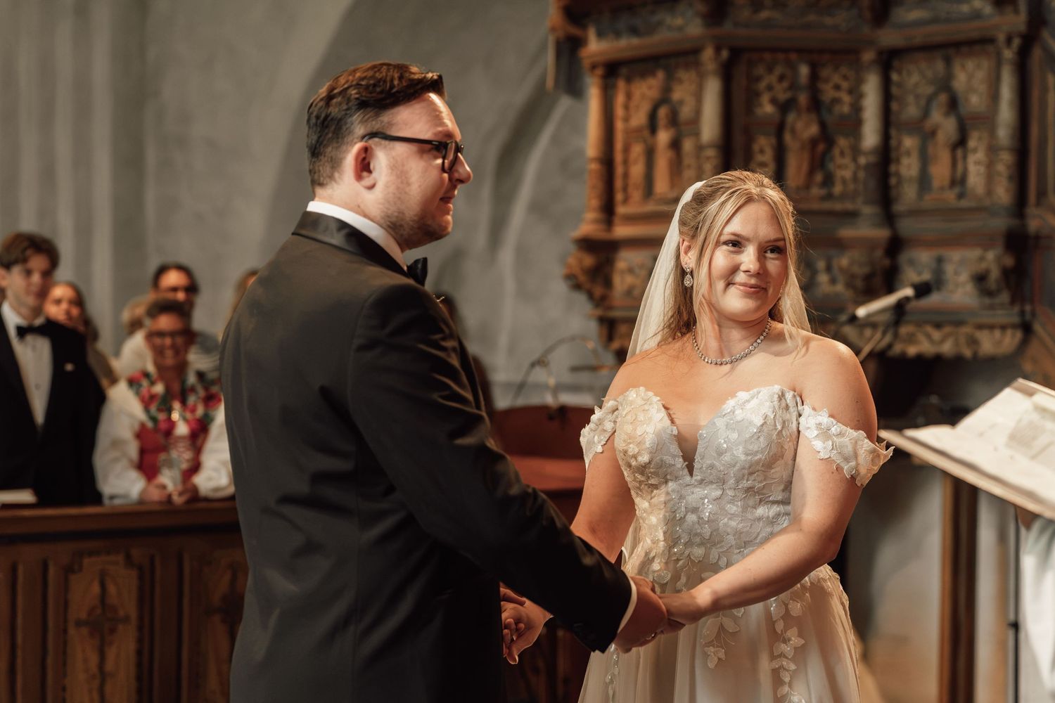 Bride and groom exchange vows at church altar, bride in lace off-shoulder gown, groom in black suit.