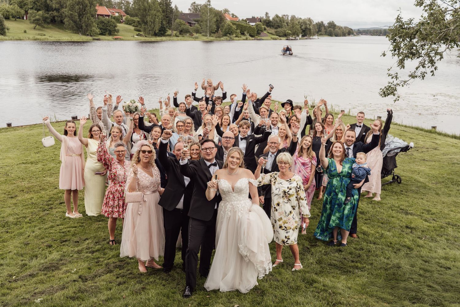 Joyful wedding group photo with bride and groom surrounded by cheering guests on a lakeside lawn.