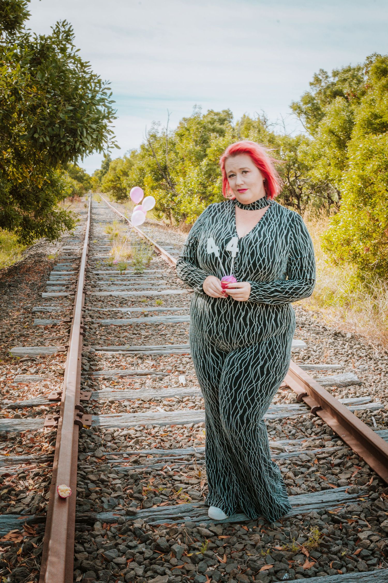 A person with bright red hair wearing a floral pattern dress poses dramatically on train tracks during an outdoor photoshoot.