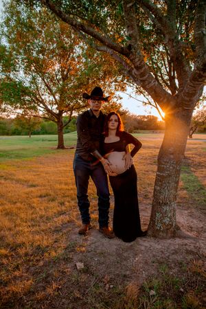 A maternity photo at sunset with a couple posing beneath a tree.