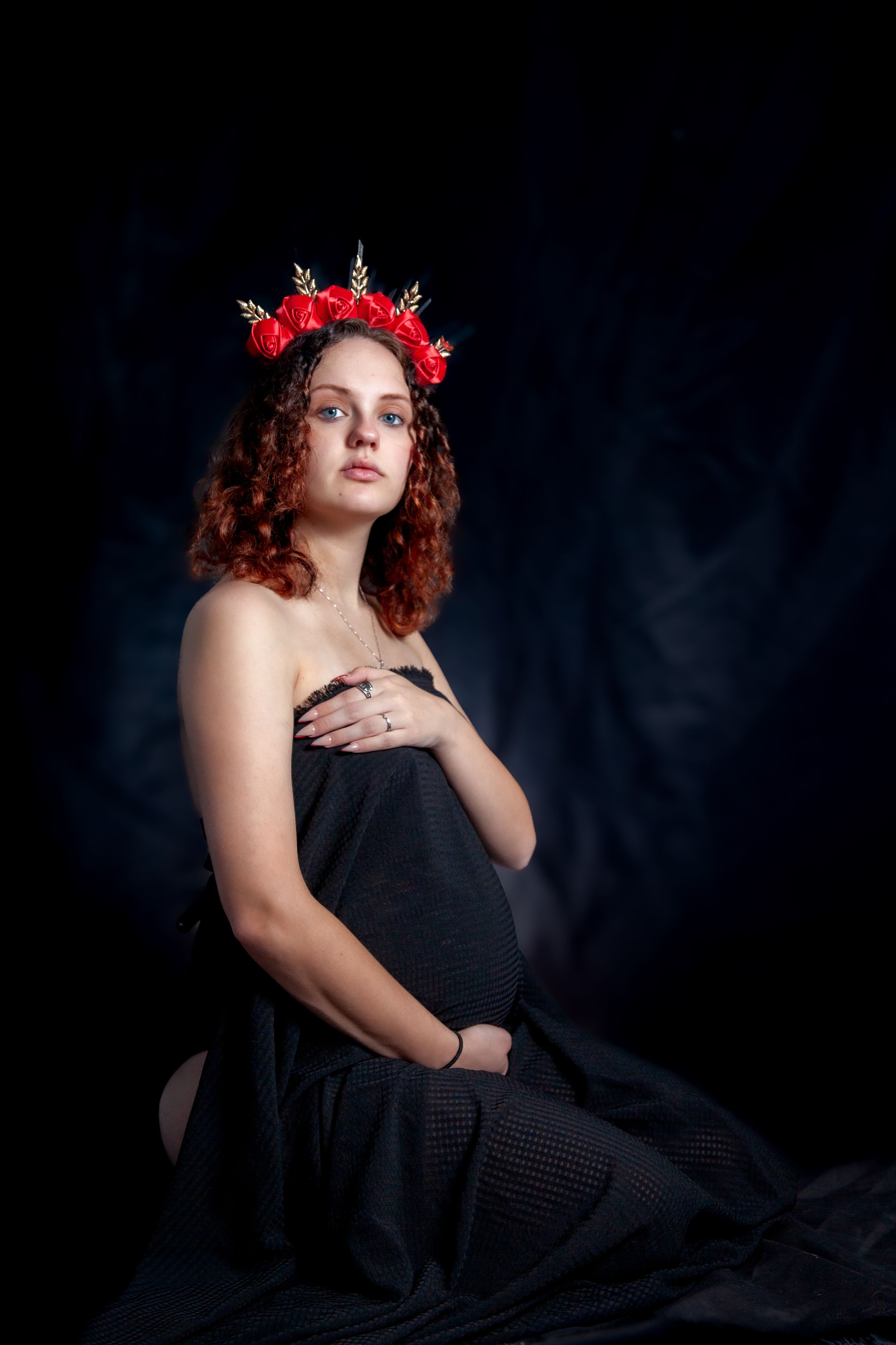 Dramatic studio portrait against dark background featuring black dress and red floral crown.