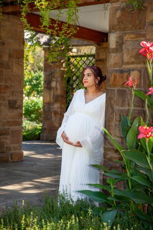A maternity portrait in a flowing white dress among blooming flowers and stone architecture.