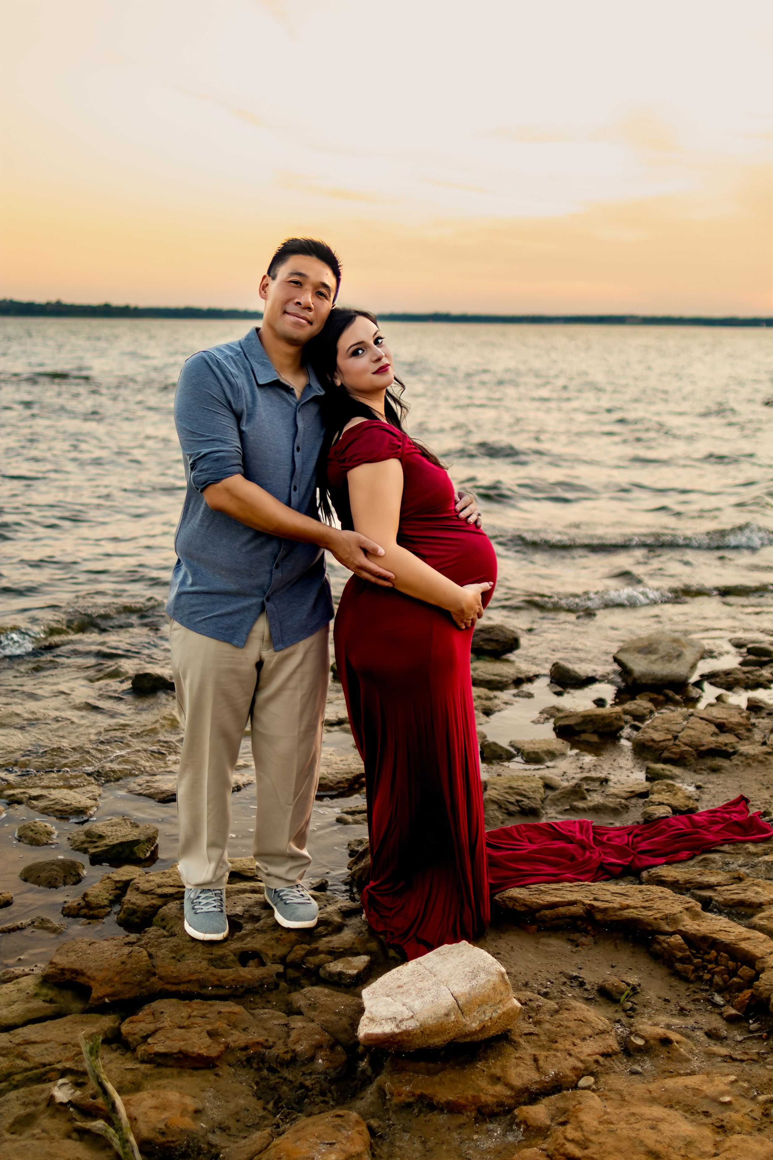 Romantic maternity photos of couple embracing on rocky shoreline at sunset.