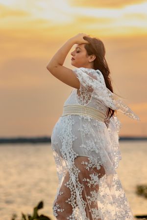 Woman in white lace dress posing dramatically against sunset over water.